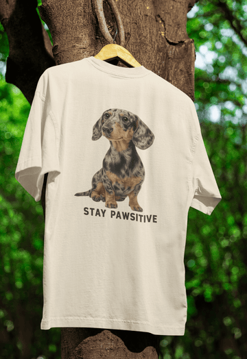 Cremefarbenes T-Shirt mit Dackel-Motiv und Schriftzug Stay-Pawsitive im realistischen Illustrationsstil für Hundeliebhaber.